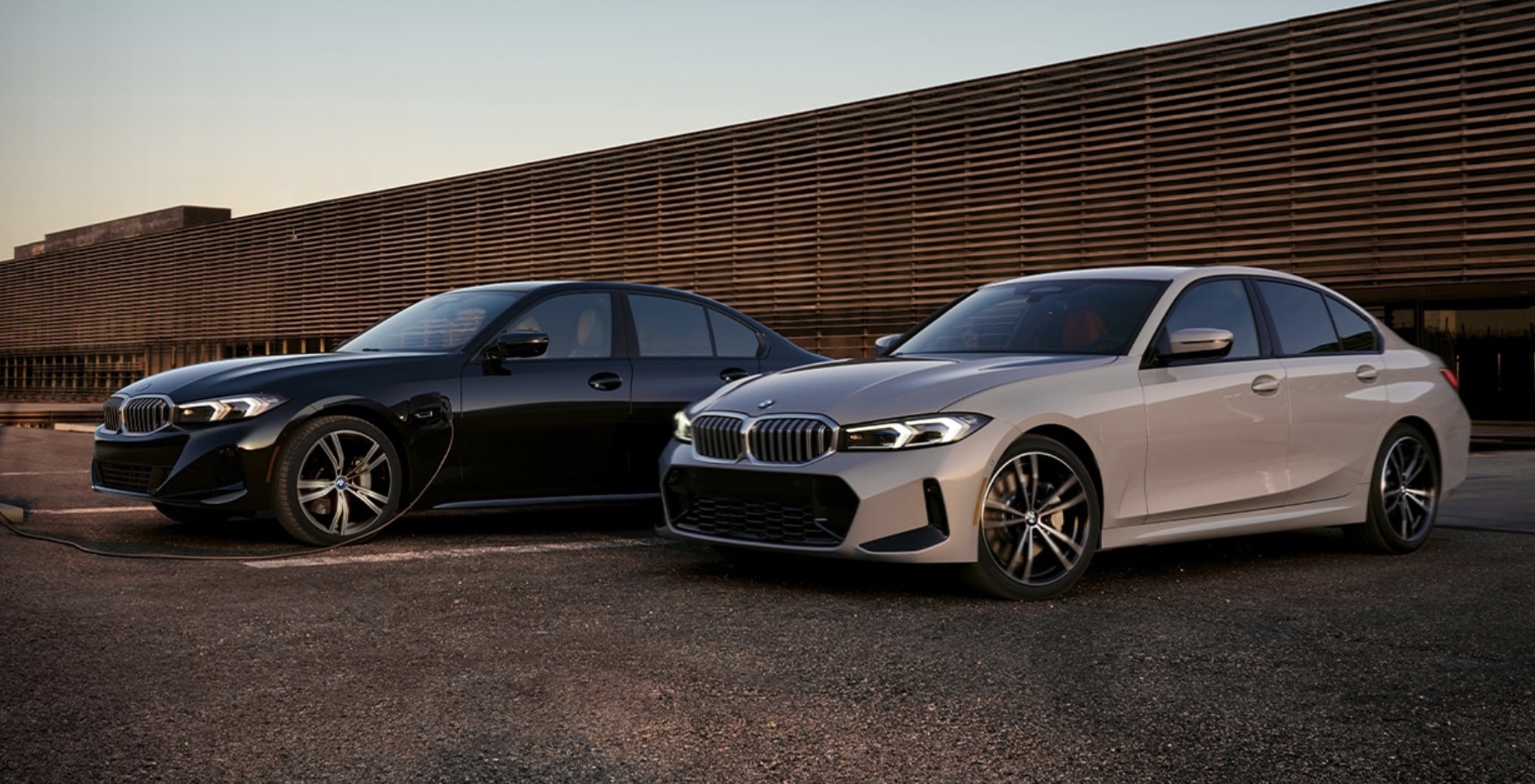 Two 2023 BMW 3 Series parked in front of an industrial building with the sun setting.